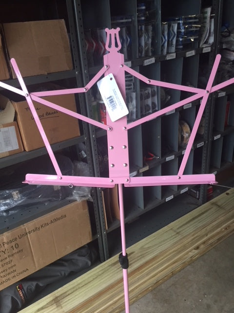 A pink aluminum music stand with a tripod base, positioned indoors, possibly in a storage area or on a stage.